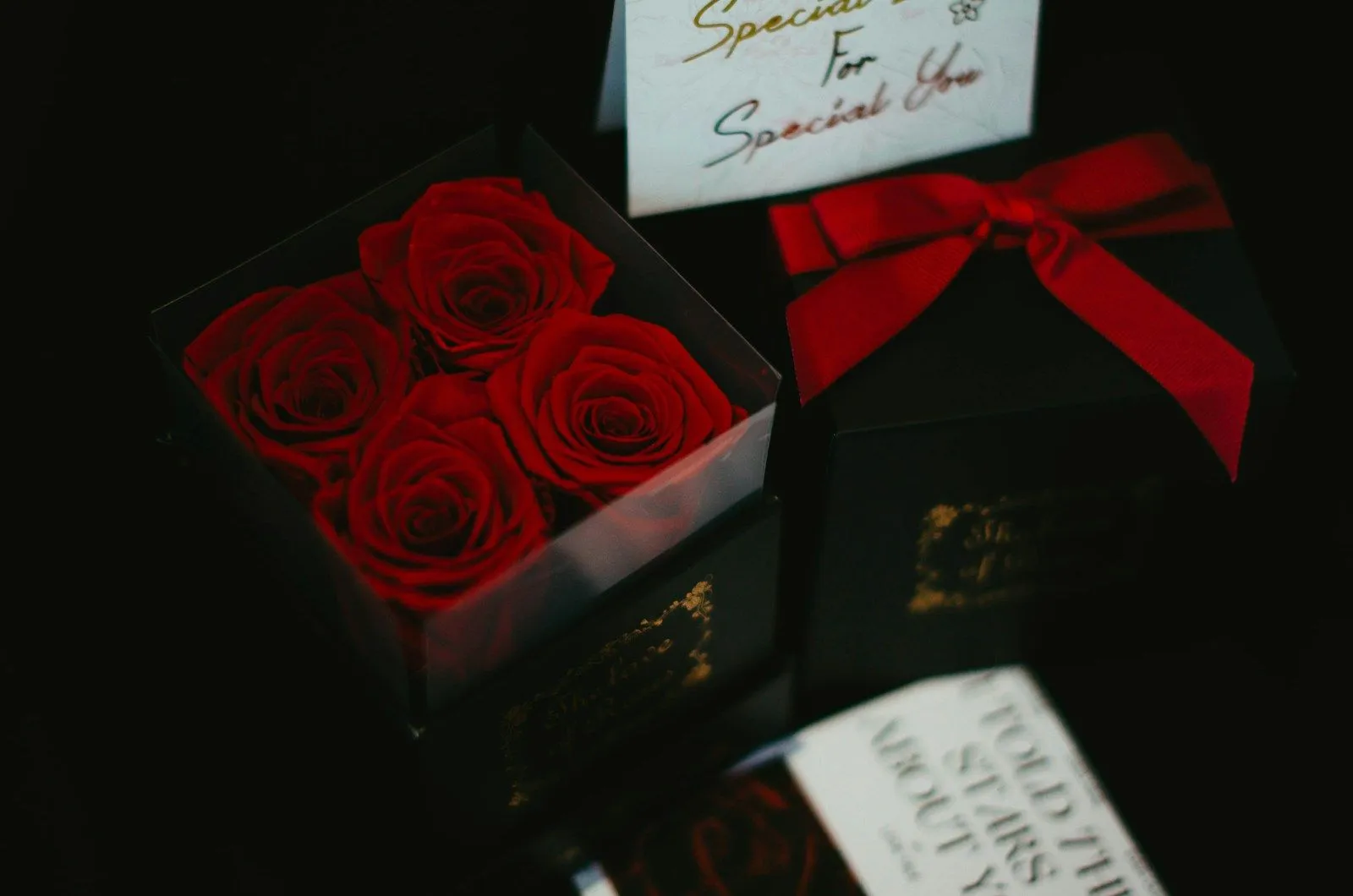 Cuatro rosas rojas preservadas dentro de una caja negra con frente transparente, junto a una caja de regalo con moño rojo